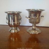 Two small silver plated urns on wooden surface showing lion head ring handles on one and leaf motif on the other