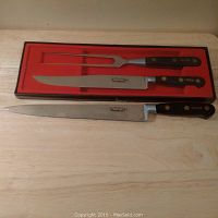 Full view of the boxed set showing two chef knives and a carving fork with black handles and brass rivets inside a red presentation box and one larger knife outside the box on a wooden surface.