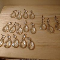 18 clear faceted teardrop-shaped chandelier crystals laid out on a light wood surface, each with original metal hooks and some with spacer crystals.