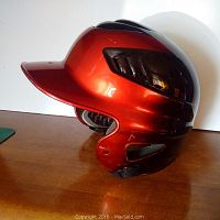 Side profile of the red and black Rawlings batting helmet on wooden surface with lighting showing gloss and vents.