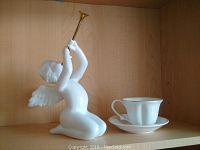 Front view of the white porcelain angel holding a gold trumpet and the heart-shaped white china cup and saucer on a wooden shelf.