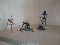 Four small metal sculptures displayed on a wooden shelf: a tripod balancing figure with brass spheres, a helicopter shape with a plier-like helicopter blade, a skier figure on a circular base, and a tall balancing figure with brass spheres on a vertical stand.