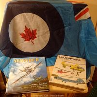Photo showing Royal Canadian Air Force Academy flag draped on a chair with two military flight books on table beneath, 'Spitfire II The Canadians' and 'The Complete Book of Helicopters'.