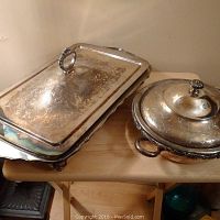 Photo showing two silver plated servers, one rectangular with a decorative ring handle and floral engraving, the other is round with cover, engraved patterns, and two side handles.