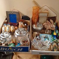 Overview of two boxes filled with various decorative items including glass jars with metal lids, wooden clogs, a rabbit figurine, metal and glass dishes, framed art, and assorted small figurines and objects.