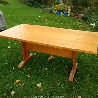 Solid wood trestle table shown outdoors on grass from an angled side view to display the tabletop and leg structure.