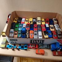 Overview of 38 metal toy cars and trucks in a cardboard box with a few lined up in front on a table showing mixed condition and wear.