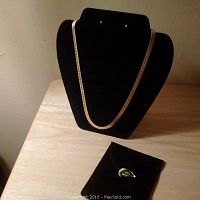 The 24 inch 14k gold plated herringbone chain necklace displayed on a black jewelry bust next to its velvet pouch.