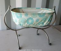 Side view of antique doll crib showing metal curved structure, small wheels, and floral plastic bassinet.