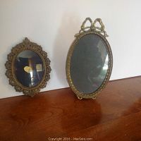 Two old-style Italian metal oval portrait frames displayed on a wooden surface against a white wall, showing condition and design details.