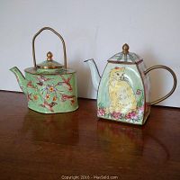 Photo showing front and side views of two miniature enamel Chinese teapots. One with hummingbird and floral decoration and the other with a cat and floral design.