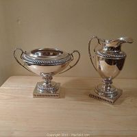 Two silver finish classical style floral containers on a wooden surface, including a covered bowl and a handled urn, both with decorative rope-like edges and square pedestal bases.