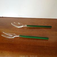 Pair of glass serving utensils with clear heads and green handles on a wooden surface.