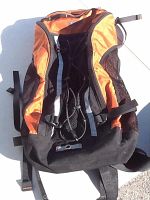 Front view of the Mountain Equipment Co-op knapsack showing orange panels and black base with adjustable straps and mesh side pockets