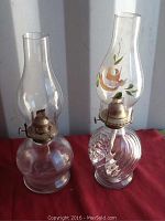 Two vintage oil lamps on red surface, showing full front view, one with plain chimney and smooth base, other with painted floral chimney and swirled base