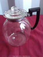 Photo showing clear glass tea pot with glass lid and black Art Deco Bakelite handle on red cloth background.