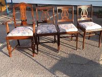 Photo showing four wooden dining chairs in a row on pavement, one armchair (left) with armrests, one fancier chair (second from left) and two common chairs. Upholstered seats have patterned fabric.