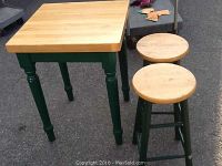 Butcher block table with two matching stools, shown outside on pavement with some items in background.