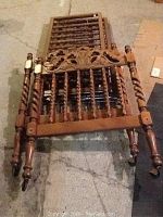 Photo shows one vintage crib end featuring turned spindles and decorative carving on top rail, wood in medium brown tone, laid flat on concrete floor.