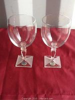 Pair of clear Bohemian crystal wine glasses with rounded bowls and square bases on a red cloth.