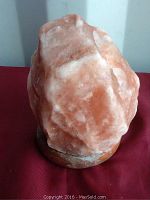 Front and side views of an approximately 6 inch tall Himalayan salt lamp mounted on circular wooden or resin stand. The lamp displays natural orange and white coloration and a rough, unpolished surface.