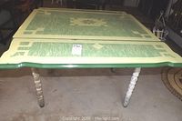 Photo showing top view of rectangular drop leaf table with enamel top, central floral geometric pattern in green and cream, minor enamel loss visible