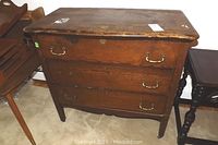 Antique solid wood dresser with shaped top and three drawers, one missing brass pull, on tapered legs with casters, visible wear on top and legs.