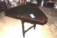Dark wood antique triangular drop leaf table with brass hinges and turned legs on carpeted floor, closed position.