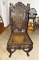 Full front view of the antique heavily carved wood chair showing the caned seat and the detailed carvings on legs, apron, and backrest with oval caned insert.