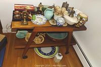 Wide shot of the wooden stand with multiple ceramic and decorative items arranged on the top and shelves below, also showing some of the wicker chairs leaning behind.