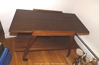 Solid teak wheeled cart seen from an angle showing rectangular solid wood top, slatted bottom shelf and angled legs with wheels.