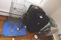 Two suitcases, one black and one blue, accompanied by a wheeled shopping cart, positioned near a wall with multiple box fans on the floor.