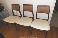 Three solid teak chairs with cream colored padded seats and caned backs, placed side by side on hardwood floor.