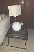 Shows black wrought iron side table with glass top alongside beige square shaded table lamp and a white decorative bulb lamp resting on the table.