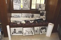 Group of ten black and white photos displayed on bench and window ledge with over 25 rolled posters placed nearby in tubes and flat arrangement