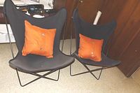 Two black wrought iron butterfly chairs with black canvas seats and orange throw pillows, facing forward.