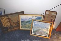 Four framed art prints resting on floor against wall, showing city street and landscape scenes.
