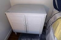 White painted wooden cabinet with two slatted doors closed, showing size and design on tapered legs.