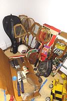 Wide shot showing multiple vintage wooden tennis rackets, badminton rackets in original packaging, a Bedford soccer ball, and weights on the floor.