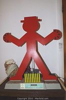 Front view of red Counting Man figure with stylized limbs and head, mounted on black base with yellow picket fence in front.