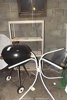 Photo showing Happy Cooker round kettle grill and round metal patio table with tubular legs and umbrella hole, along with plastic shelving unit in background