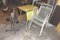 Photo of yellow laminate drop leaf table with metal trim and four tapered legs alongside three white painted metal wired chairs and a black vinyl seat exercise ergometer.