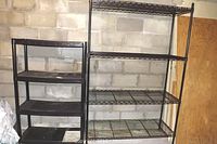 Pair of shelving racks positioned against a cinder block wall: smaller black plastic rack on the left, taller black metal wire rack on the right.