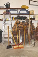 Overall lot showing metal ladder, firewood stack, garden tools including shovels, rake, saws and broom