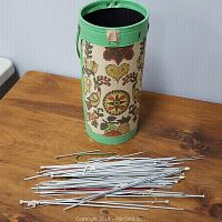 Full view of assorted knitting needles lying next to upright decorative cylindrical holder with floral fabric and green trim.