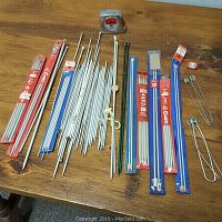 Full lot shot showing knitting needles in packages and loose, metal stitch holders, measuring tape on wooden table.