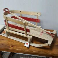 Wooden sleigh placed on wooden table showing front and side view with red accents and pull handle with red grip.