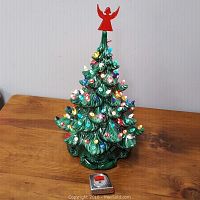 Full front view of the green ceramic Christmas tree decorated with multi-colored bulbs and red angel topper, sized next to a tape measure.