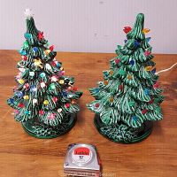 Two green ceramic Christmas trees with multi-colored bulbs on a wooden surface with a 12 inch tape measure for scale, showing size and condition