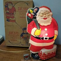 Plastic illuminated Santa Claus figure in red suit holding a green toy sack, with original cardboard box behind it featuring product details and illustrations.